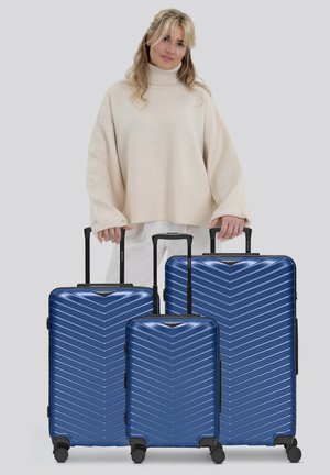 Donna in maglione crema che tiene le maniglie di tre valigie rigide blu con ruote, disposte per dimensione davanti a lei su uno sfondo grigio.