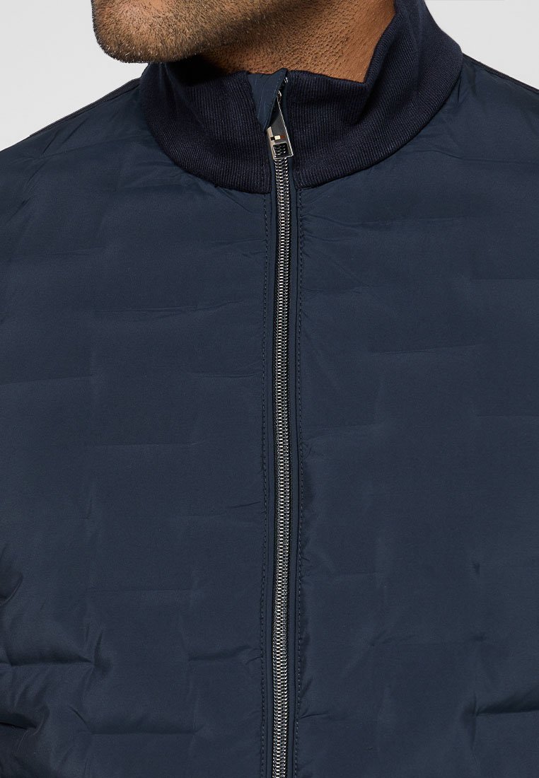 Giacca puffa blu navy con colletto alto, texture trapuntata e chiusura frontale con zip. Il materiale appare leggero e isolato.