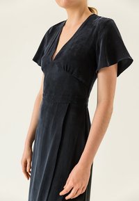 Černé šaty s výstřihem do V, krátkými rukávy, měkkou texturou, přiléhavým trupem a splývavou sukní; obsahují jemné plisování a minimální designové detaily.