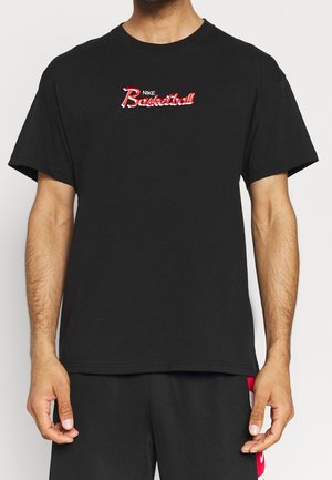 Czarna koszulka z krótkim rękawem z czerwonymi i białymi napisami "Nike Basketball" na piersi, noszona przez osobę na jasnym, jednolitym tle.