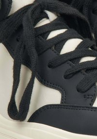 Sneaker wysoko cholewkowy z czarną skórzaną cholewką, z kremowymi akcentami, grubymi czarnymi sznurowadłami i teksturowanymi powierzchniami. Posiada kontrastowe szwy.
