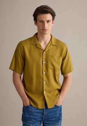 Joven con una camisa de botones de manga corta color mostaza y vaqueros azules, de pie con las manos en los bolsillos delante de un fondo liso.