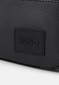 Czarna torba z perforowanej skóry, charakteryzująca się teksturowaną powierzchnią oraz prostokątną naszywką z logo "HUGO" wytłoczonym na czarno.