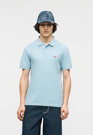 Jeune homme portant un polo bleu clair, un pantalon bleu foncé à fines rayures et une casquette de baseball bleue à motifs, debout devant un fond uni.