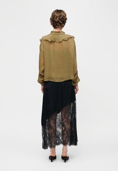 Άτομο που φοράει ελαιδί διάφανη μπλούζα με γιακά με βολάν και μαύρη ασύμμετρη φούστα με διάφανο δαντελένιο τμήμα, στέκεται με την πλάτη προς τον θεατή.