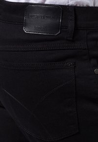 Jeans de mezclilla negra con una etiqueta de cuero que presenta "MASTERPIECE" y "BRAX FEEL GOOD." Incluye detalles de costura en los bolsillos y las costuras.