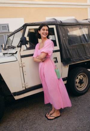 Ung kvinde i pink kjole smiler med korslagte arme, stående ved siden af et hvidt vintage terrængående køretøj på en bygade.