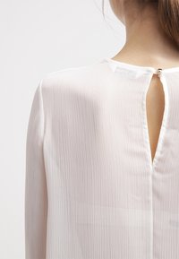 Camicia bianca leggera e trasparente con una superficie morbida e testurizzata. Presenta una chiusura a occhio di serratura sulla schiena fissata con un piccolo bottone.