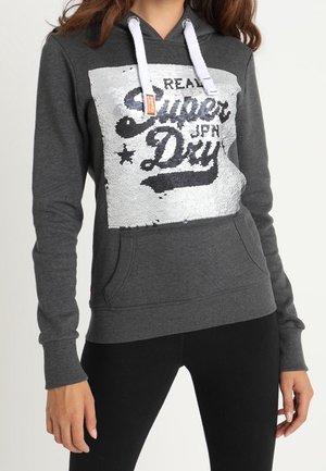 Femme portant un sweat à capuche gris foncé avec des cordons blancs et un patch à sequins indiquant "Real Superdry JPN Day" et un pantalon noir.