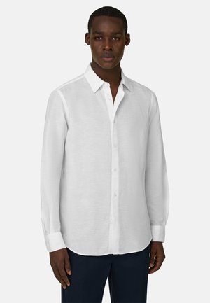 Hombre con una camisa blanca sencilla de manga larga abotonada y pantalones oscuros, de pie frente a un fondo claro liso.