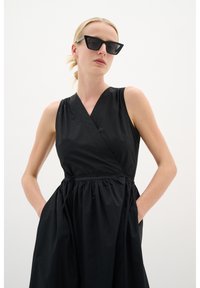 Schwarzes ärmelloses Kleid mit Wickeldesign, gerafftem Rock und seitlichen Taschen. Glatte Textur mit minimalistischem Ästhetik. Schwarze Sonnenbrille getragen.