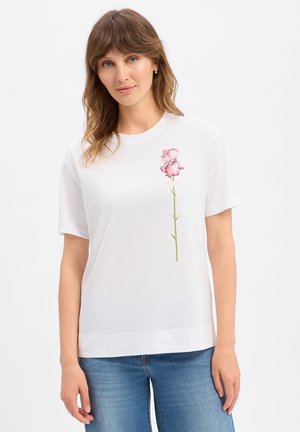 Vrouw met lichtbruin haar die een wit T-shirt met een roze bloemafbeelding en blauwe spijkerbroek draagt tegen een effen witte achtergrond.
