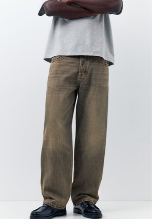 Person med krydsede arme iført en grå skjorte, løse brune jeans og sorte polerede loafers, der står mod en ensfarvet baggrund.