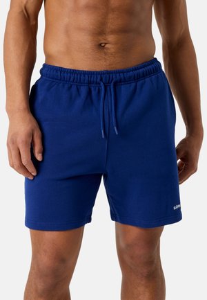 Torse masculin portant un short de sport bleu à cordon, debout avec les bras détendus le long du corps sur fond blanc.