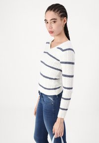 Suéter blanco de punto de cable con rayas azul marino, diseño de cuello en V y mangas largas, combinado con jeans azul oscuro y un bolso azul claro.