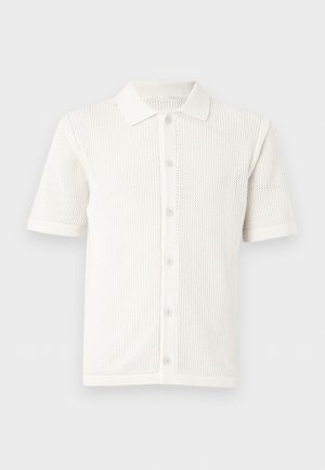 Młody mężczyzna z kręconymi włosami, ubrany w kremową koszulę i beżowe szerokie spodnie, trzyma złożoną brązową skórzaną torbę na białym tle.