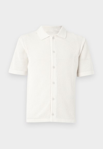 Λευκό πλεκτό πόλο με κοντά μανίκια, κουμπιά μπροστά και υφασμένο ανοιχτό σχέδιο σε όλη την επιφάνεια.