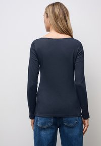 Marineblaues Langarmoberteil mit weit ausgeschnittenem Kragen, weichem Material und figurbetonendem Design. Kombiniert mit blauen Jeans, die Rückendetails zeigen.