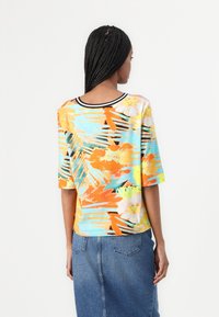 Kurzärmliges T-Shirt mit Rundhalsausschnitt, das ein lebhaftes, mehrfarbiges Blumen- und Tropenmuster auf einem hellblauen Hintergrund zeigt. Schwarze Einfassung am Ausschnitt.