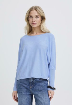 Donna bionda che indossa una maglia a maniche lunghe di colore azzurro chiaro e ampia, abbinata a jeans blu e un braccialetto nero, in piedi davanti a uno sfondo uniforme.