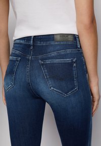 Jeans ajustados en denim azul oscuro, con un pequeño logo en la cintura, dos bolsillos traseros con detalles de costura y un leve efecto de desgaste.