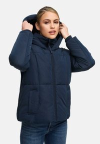 Vrouw die de capuchon van een marineblauwe pufferjack aanpast, gekleed in donkere jeans, staande tegen een effen lichte achtergrond.