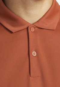 Polo couleur rouille présentant une texture lisse, un col classique et une patte de boutonnage à deux boutons de la même teinte. Convient pour une tenue décontractée.