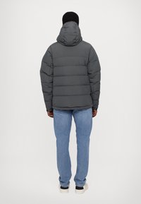 Veste matelassée grise avec capuche, texture capitonnée et poignets élastiques, portée avec un jean bleu clair et des baskets blanches, vue de dos.