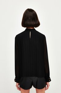 Blusa nera plissettata con maniche lunghe, colletto alto, dettaglio keyhole sul retro e rifiniture con bottoni dorati ai polsini. Abbinata a pantaloni corti neri.