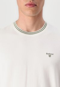 Tricou alb din bumbac cu guler ribbed în dungi verzi și albe, având un mic logo Brodat Barbour pe partea stângă a pieptului.