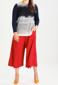 Laivansininen ja harmaa pitkähihainen paita abstraktilla kuviolla, yhdistettynä punaisiin leveälahkeisiin housuihin. Mallilla on päällään keltaiset korkokengät.