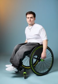 Manuaalratastool mustade rataste ja roheliste aktsentidega, hallid laiade jalgadega teksad, valge lühikeste varrukate särgid ja valged spordijalatsid.