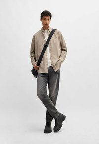 Bruine button-up shirt over een witte T-shirt, grijze jeans en zwarte veterschoenen. De model staat met een zwarte schoudertas voor een neutrale achtergrond.