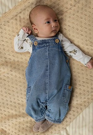 Salopette en denim bleu clair avec boutons en bois et poches latérales, portée sur une chemise crème à imprimés animaux, sur un fond de couverture texturée.