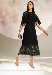 Fekete estélyi ruha feszített felsőrésszel, bővülő szoknyával és díszített szegéllyel. Átlátszó, hímzett köpenyujjak adnak textúrát és részletességet. Ezüst magassarkú cipők.