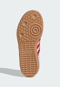 Adidas obuća s gumenom gumom koja ima teksturirani uzorak, crvene naglaske na bijelom gornjištu i utisnuti logo.