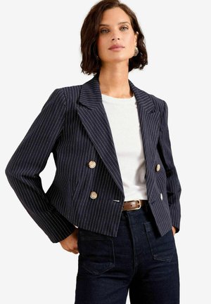 Tummansininen raidoitettu blazer, jossa on kaksoisnapit, lovet ja lyhyt malli, yhdistetty valkoiseen t-paitaan ja tummiin farkkuihin.