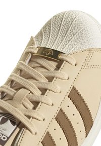Beige Adidas sneaker close-up showing textured shell toe, cream laces with gold metal lace tag, and brown stitched three stripes.
