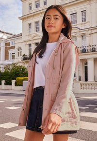 Chaqueta impermeable de color rosa claro con capucha, cierre de cremallera en la parte delantera y cintura elástica, llevada sobre una camisa blanca y una falda de mezclilla oscura.
