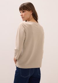 Frau mit braunem Haar, die ein beige-weiß gestreiftes Langarmshirt und dunkelblaue Jeans trägt, leicht zur Seite gedreht.