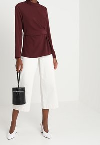Maglietta bordeaux a maniche lunghe con collo alto e drappeggio davanti, abbinata a pantaloni bianchi a gamba larga e borsa nera dal finish texture. Tacchi bianchi.