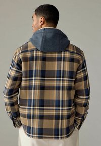 Bruine en marineblauwe geruite blouse met een grijze capuchon, voorzien van een zachte textuur en een relaxte pasvorm. De blouse heeft lange mouwen en een rechte zoom.