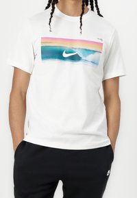 Bílý bavlněný tričko s barevným surfovým motivem, zobrazující modrou oceán, vlny a růžový západ slunce, doplněný středovým logem Nike.