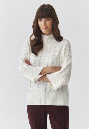 White cable-knit sweater with a high neck and 3/4 sleeves, featuring ribbed cuffs and hem. Paired with deep burgundy trousers.