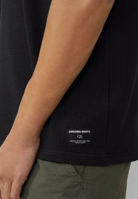 Zwart gestructureerd shirt met korte mouwen met een zichtbaar label "EVOLVING ROOTS" aan de onderkant, gecombineerd met groene broek.