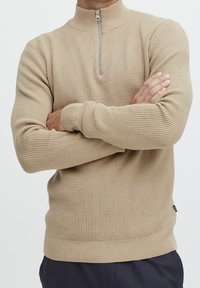 Beżowy sweter z długim rękawem, z wysokim kołnierzem i zamkiem do ćwiartki. Teksturowana, dzianinowa tkanina z prążkowanymi mankietami i dole, dopasowany fason.