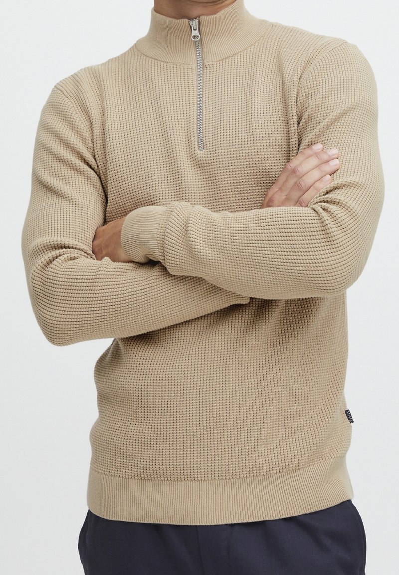 Beżowy sweter z długim rękawem, z wysokim kołnierzem i zamkiem do ćwiartki. Teksturowana, dzianinowa tkanina z prążkowanymi mankietami i dole, dopasowany fason.