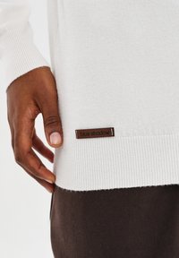 Ciemnoskóra ręka dotykająca dolnej krawędzi białego swetra z małą brązową metką "niebieski cień", w połączeniu z ciemnobrązowymi spodniami.