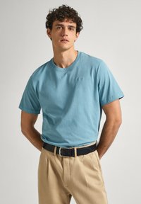 Jasnoniebieska bawełniana koszulka z krótkim rękawem, okrągłym dekoltem, z delikatnym logo. W zestawieniu z beżowymi spodniami i czarnym paskiem.