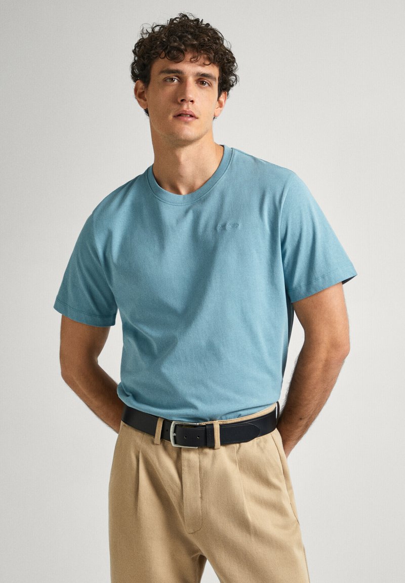 Jasnoniebieska bawełniana koszulka z krótkim rękawem, okrągłym dekoltem, z delikatnym logo. W zestawieniu z beżowymi spodniami i czarnym paskiem.